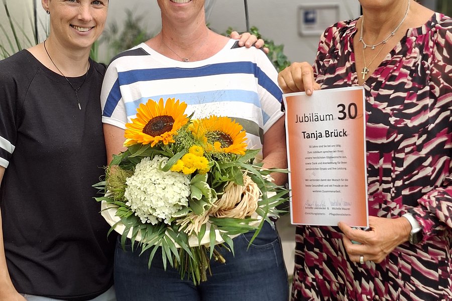 Drei Frauen stehen nebeneinander, die mittlere hält einen Blumenstrauß, die rechte eine Urkunde mit der Überschrift 'Jubiläum 30 Tanja Brück'