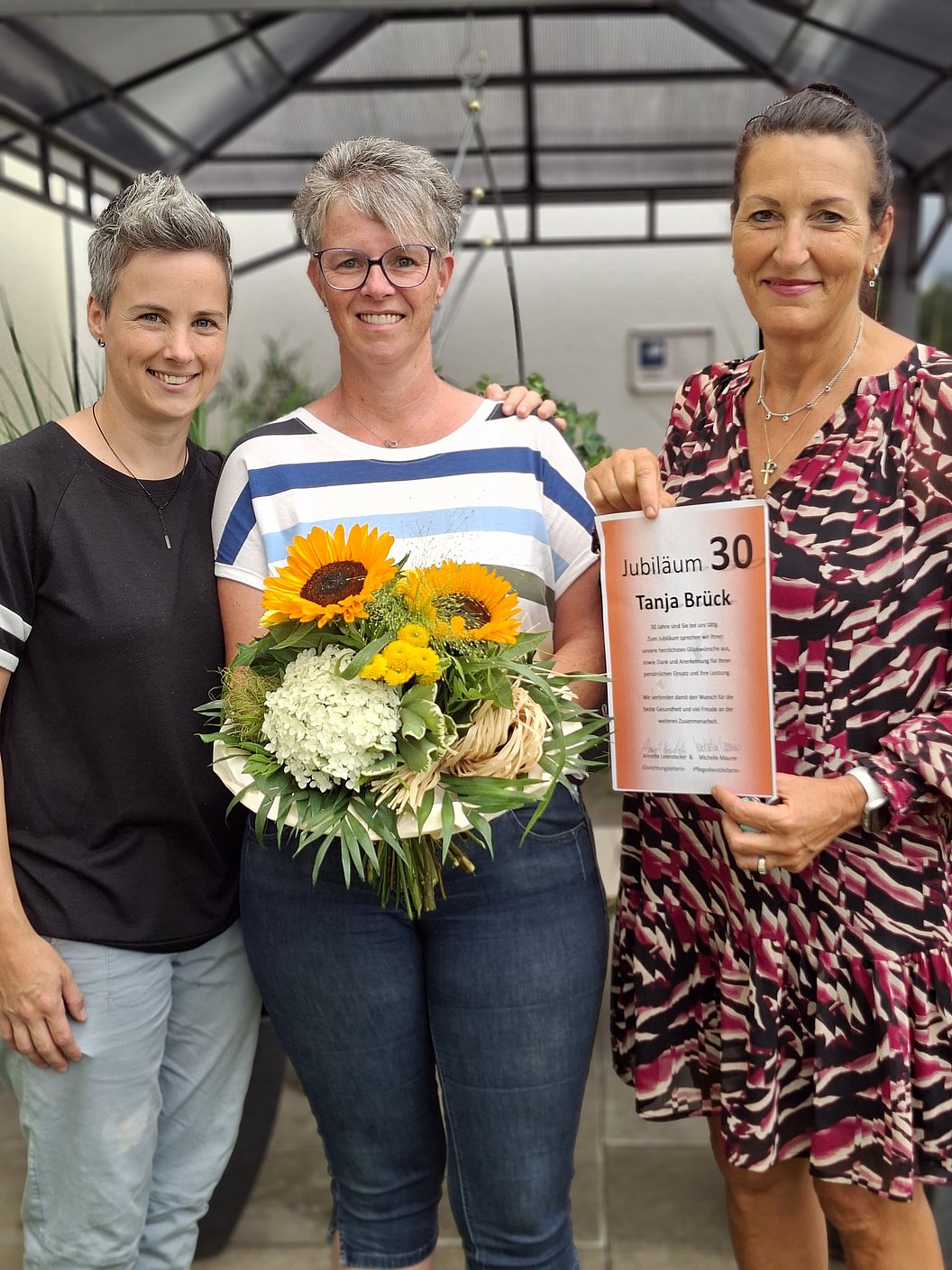 Drei Frauen stehen nebeneinander, die mittlere hält einen Blumenstrauß, die rechte eine Urkunde mit der Überschrift 'Jubiläum 30 Tanja Brück'