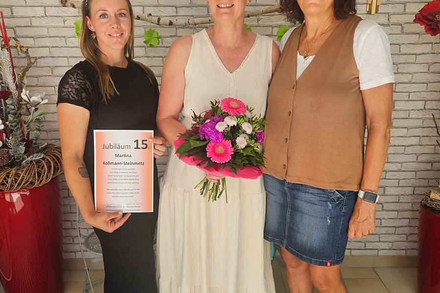 Drei Frauen stehen nebeneinander vor einer weißen Ziegelwand, die mittlere trägt ein weißes Kleid und hält einen Blumenstrauß, die linke hält eine Urkunde.