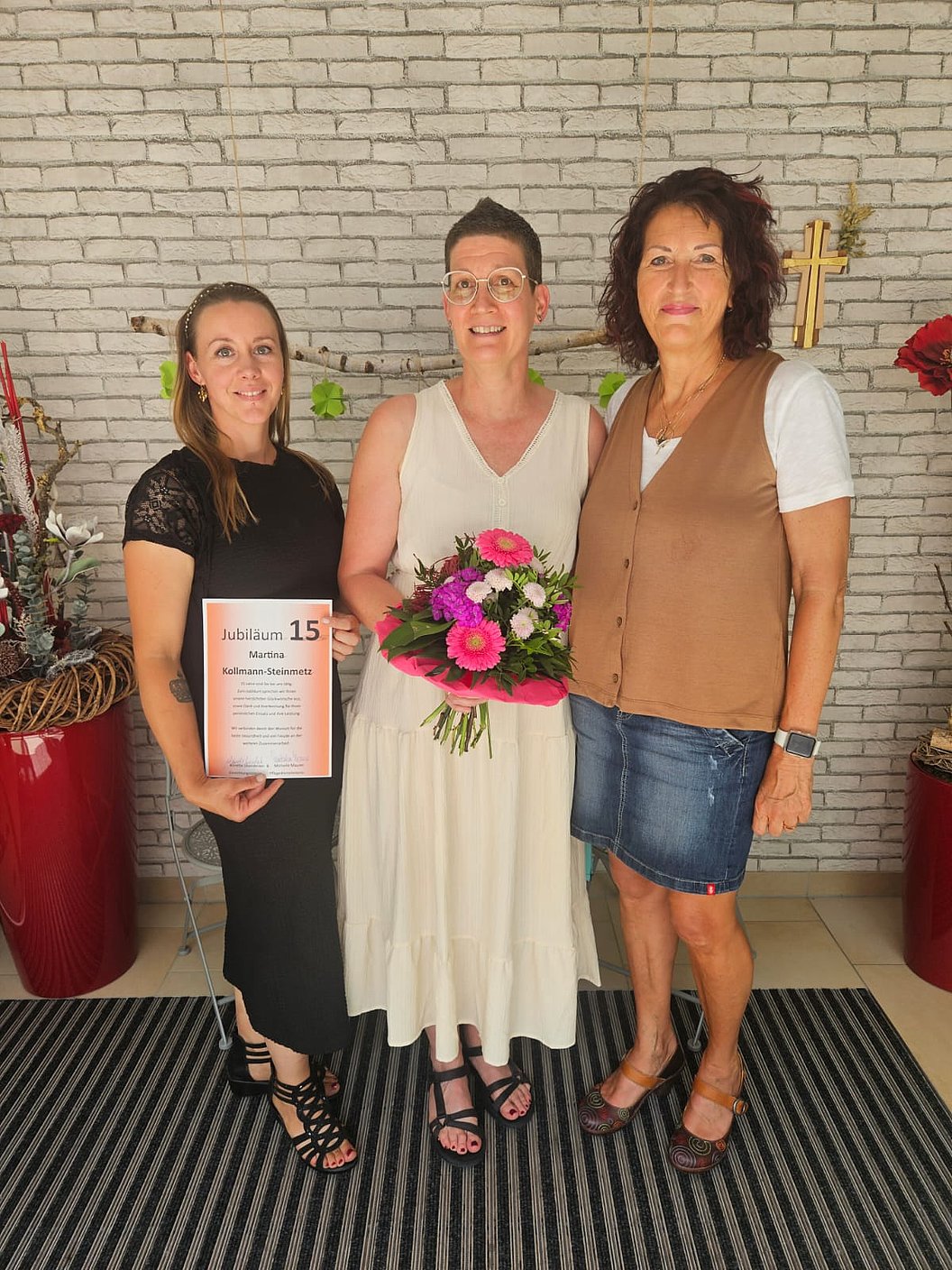 Drei Frauen stehen nebeneinander vor einer weißen Ziegelwand, die mittlere trägt ein weißes Kleid und hält einen Blumenstrauß, die linke hält eine Urkunde.